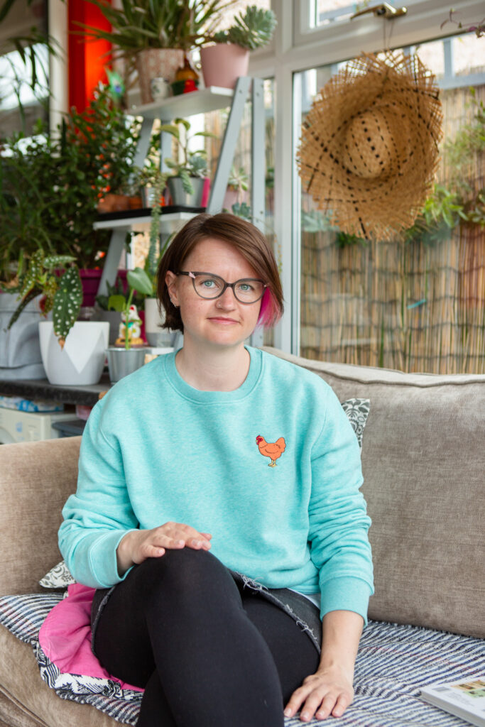 Photographic portrait of a person with short brown and pink hair, wearing cat-eye glasses, a turquoise sweatshirt with a chicken embroidered on it, black tights and a denim skirt. They are sitting on a striped blanket on a sofa, looking into the camera. They are in a conservatory with potted plants on shelves behind, a straw hat hanging from a window to the right with bamboo fencing visible through the window.