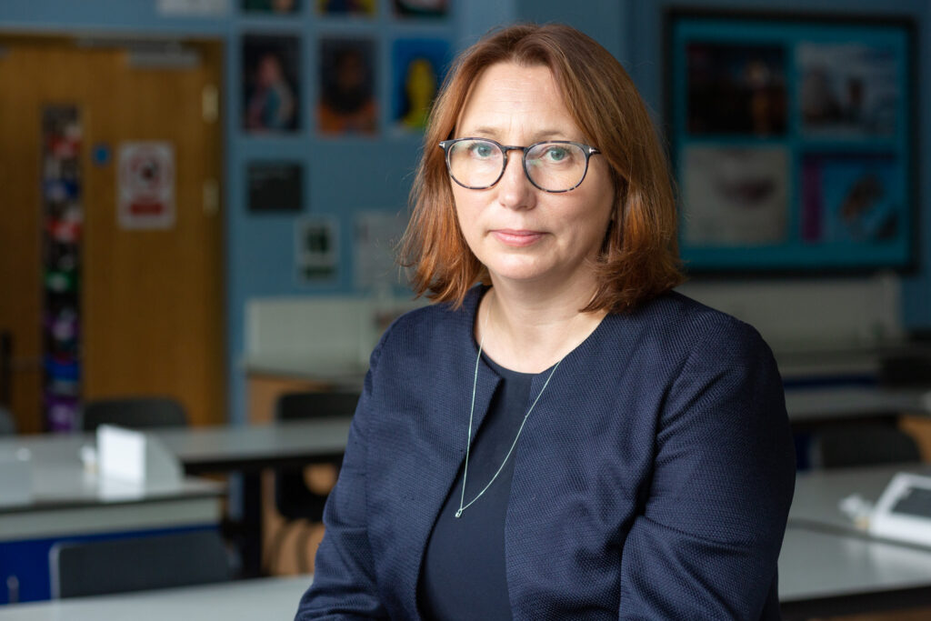 Photograph of a woman wearing glasses, with shoulder length light brown hair, dark blue suit and silver necklace, looking into camera. Behind and out of focus behind is a classroom with desks and wall displays.