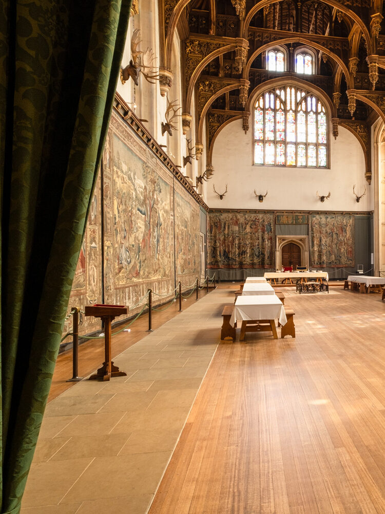 Photograph in Hampton Court Palace looking through two green curtains that are tied back either side of the frame, into a large room with wooden floor, tapestries and stag heads lining the walls, banquet tables with white table cloths and wooden stools around the edges, ornate carved wooden ceiling and an arched doorway at the far end.