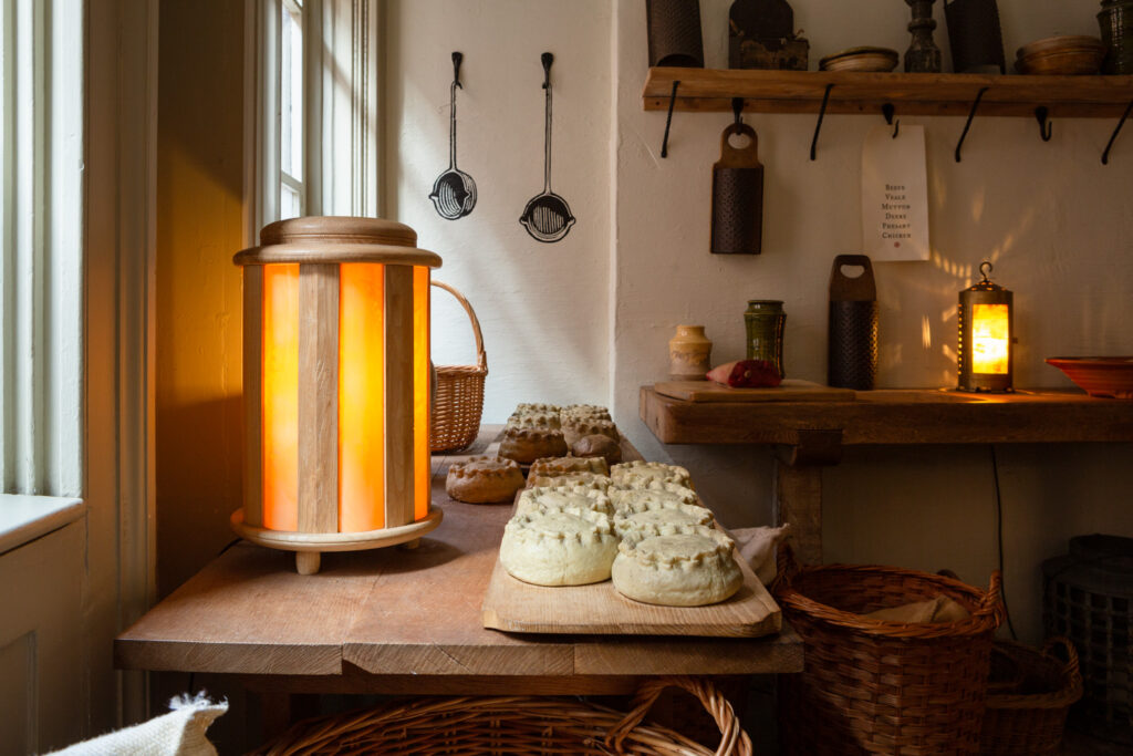 Photograph of a wooden table in front of two windows, topped with a glowing wooden lamp, replica pies on top and a basket behind. In the background is a wall with illustrations of two long-handled pans painted on the white wall and shelves to the right lined with kitchen objects and a glowing lantern.