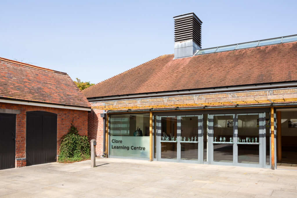 Hampton Court Palace. Photograph by Jayne Lloyd for Historic Royal Palaces. Photograph of the Clore Learning Centre at Hampton Court Palace, a brick building with tiled roof, a large metal chimney-like element on the top, glass doors with little figurines along the centre, 'Clore Learning Centre' etched in the glass and a stone paved floor in the foreground with a barn-like building to the left.