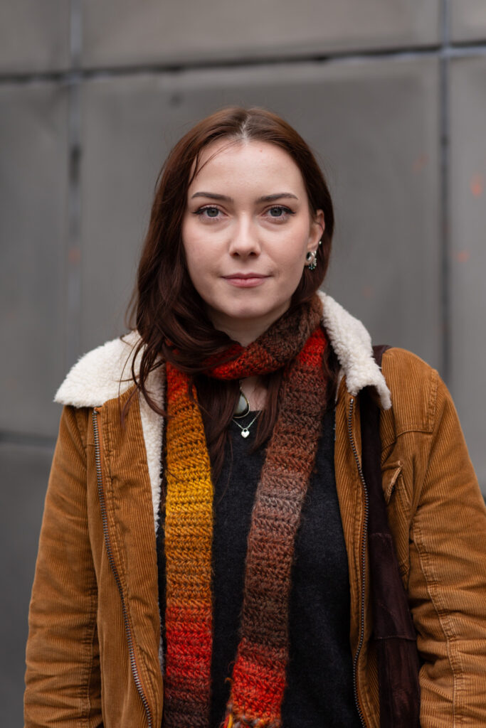 Photographic portrait of a person with dark brown hair looking into camera, wearing a thin knitted scarf in shades of orange and brown, a light brown cordoroy jacket with white collar and a dark top. They are framed by the lines of the Faraday Memorial behind.