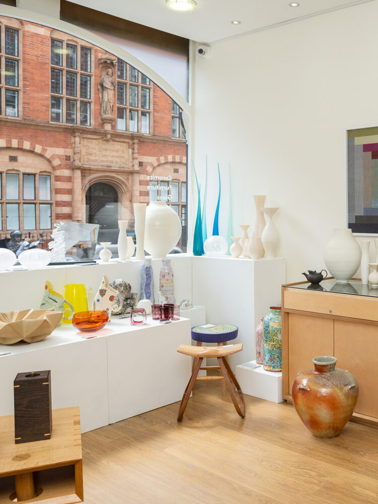 Photograph in a gallery space filled with ceramics, glass and furniture displayed on plinths and display cases and textiles mounted on the wall. A red-brick ornate building is visible through an arched window to the left of the frame.