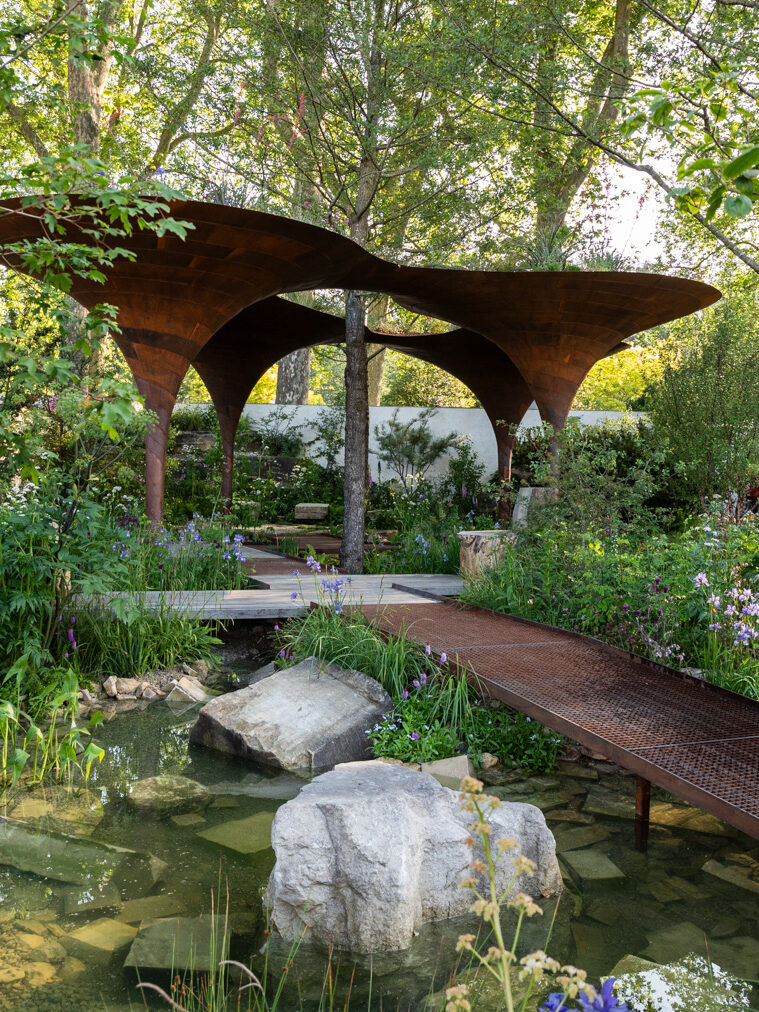 Photograph of a garden with large rusty-metal structures surrounding a tree towards the back centre of the frame, with a raised metal walkway leading towards it from the bottom right of frame. Lush green plants and flowers surround these, and in the foreground is a pool of water with stones and blocks of concrete in.