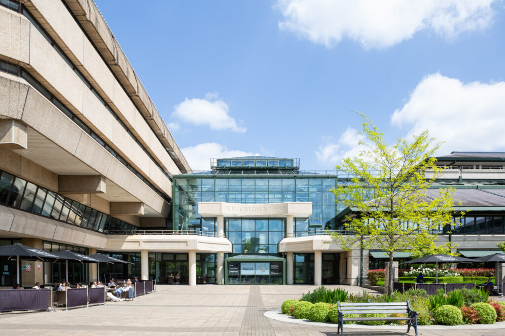 The National Archives: Spirit of Invention. Photograph by Jayne Lloyd for The National Archives. Photograph of the exterior of The National Archives building in Kew, with 'Public Record Office' inscribed above the entrance, blue sky and clouds above and trees and plants with a bench in the foreground to the right and people sitting under umbrellas to the left.