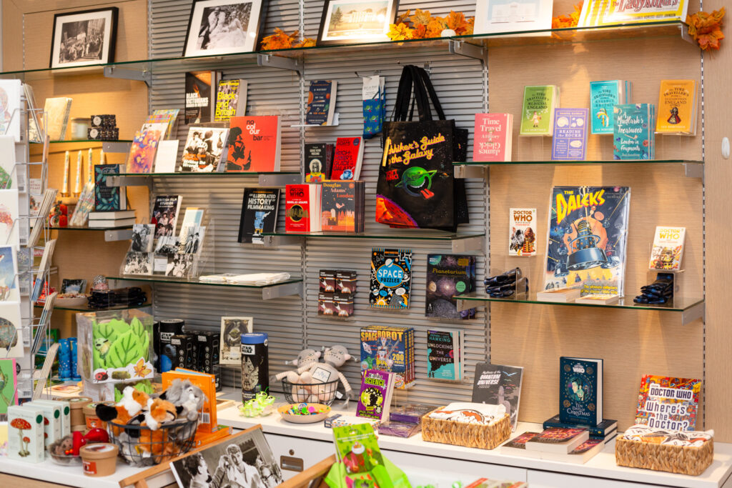 Set to Stun at Gunnersbury Park Museum. Photograph by Jayne Lloyd. Photograph of the shop at Gunnersbury Park Museum, showing items for sale including books, tote bags and toys all displayed on shelves along the wall.