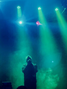 Photograph of a man singing into a microphone, with green lights shining down above, lighting a cloud of smoke behind him.