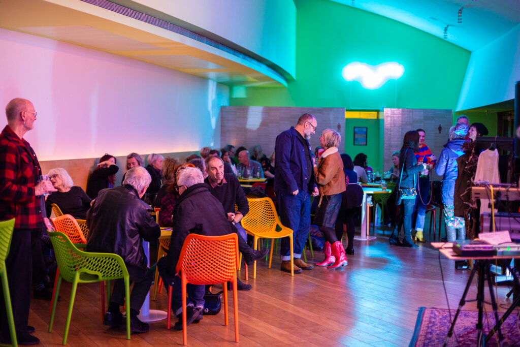 Late Lounge event at Firstsite, Colchester. Photograph by Jayne Lloyd. Photograph of the Firstsite café, at night, with people sitting in subdued colourful light at tables with colourful chairs. In the centre two people are talking and laughing and on the far wall above the entrance to the toilets a neon sign is visible but overexposed.