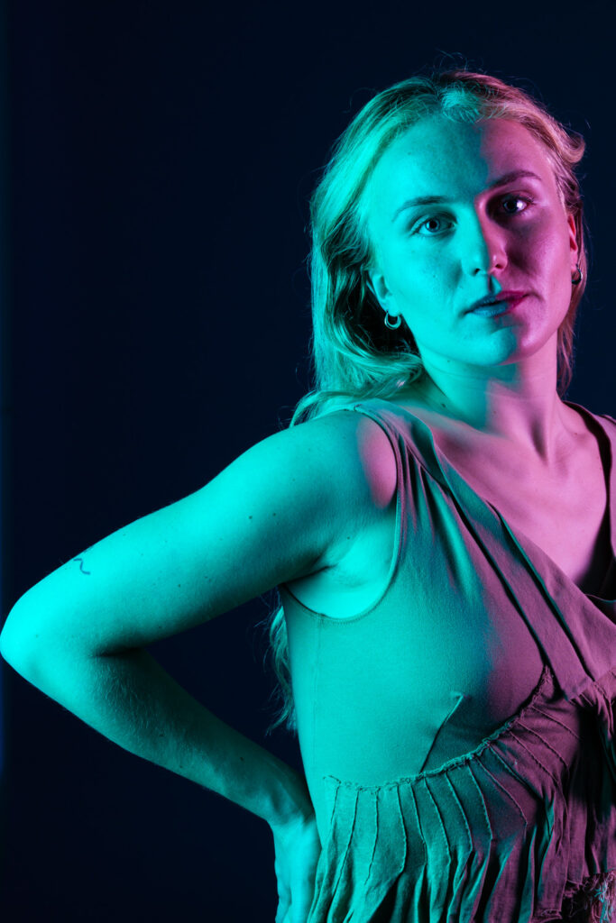 Photograph of a person with long blonde hair, framed to the right of the frame, looking into the camera and with one arm up on their hip. They are lit with blue turquoise light to the left and pink light to the right.