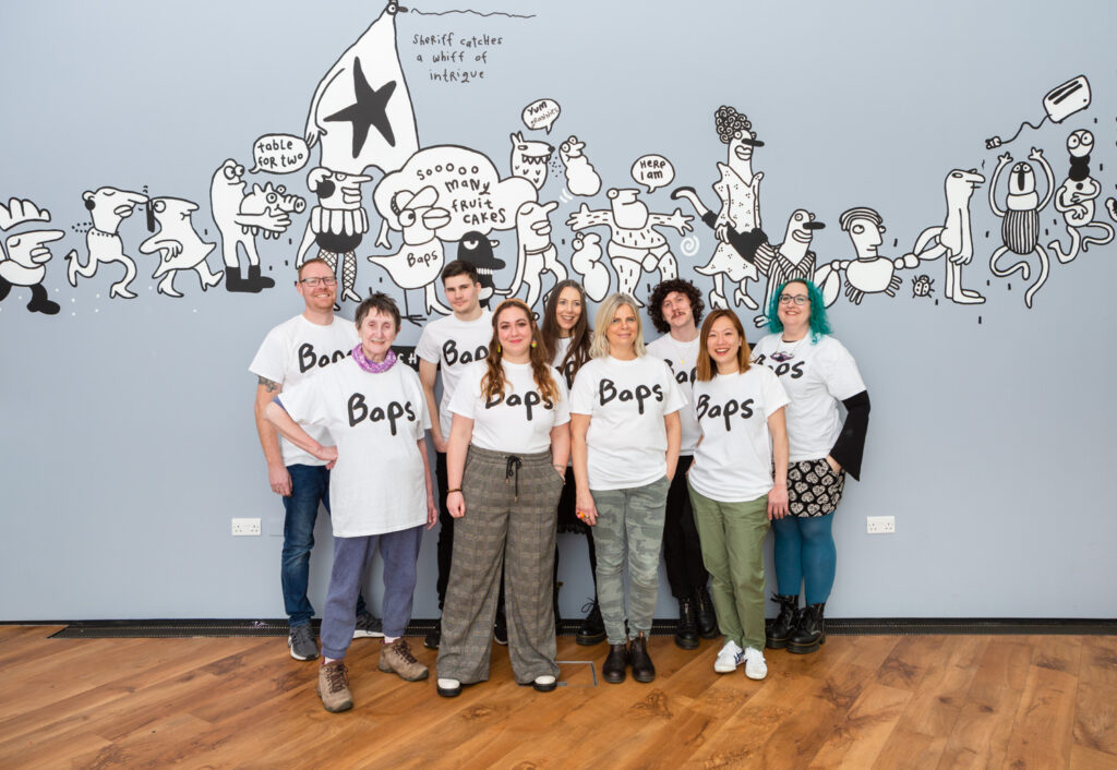 Baps T-shirt by Erica Åkerlund, available at Firstsite. Photograph by Jayne Lloyd. Photograph of 9 people modelling Baps T-shirts by Erica Åkerlund, with artwork by Erica Åkerlund on the wall behind.