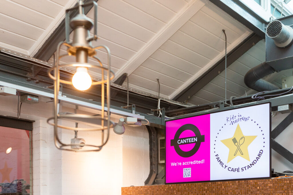 Kids in Museums Family Café Standard Launch at the London Transport Museum. Photograph by Jayne Lloyd for Kids in Museums. Kids in Museums Family Café Standard Launch, showing a digital screen showing the CANTEEN logo, 'We're accredited!', a QR code on pink to the left and 'Kids in Museums', a yellow star with a crossed fork and spoon in the centre and 'FAMILY CAFÉ STANDARD' to the right. above is the roof of the building and in the foreground is an industrial style lantern.