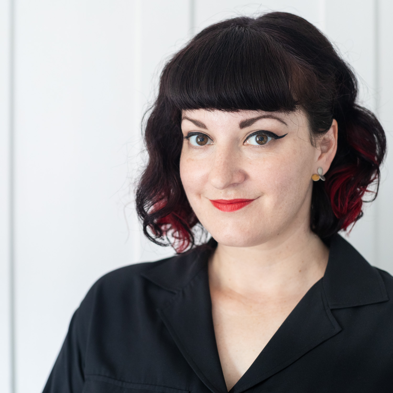 Jayne Lloyd (self-portrait) Photograph of Jayne, a white woman against a wooden panelled wall painted white. Jayne has chin-length wavy black hair with a fringe and bright red streaks underneath to each side, black winged eyeliner, pale skin and red lipstick. She is wearing a black top.